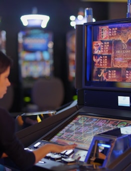 A person is seated at a slot machine console in a casino, focusing intently on the game displayed on the screen. The background shows several other illuminated gaming machines, adding a vibrant and colorful atmosphere to the scene.
